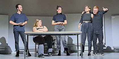 Auf dem Foto ist das Ensemble des Improvisationstheater zu sehen. Insgesamt fünf Menschen stehen auf einer Bühne. Sie alle tragen schwarze Klamotten.