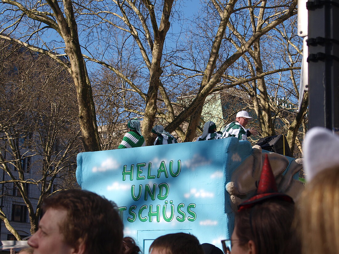 Das Hinterteil eines Mottowagens auf welchen in grün auf blauem Hintergund "Helau und Tschüss" geschrieben steht.