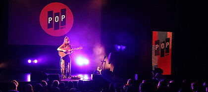 Zu sehen ist Philine Sonny während ihres Auftritts. Sie spielt Gitarre und singt auf der Bühne, im Hintergrund sind blaue Lichter zu sehen.