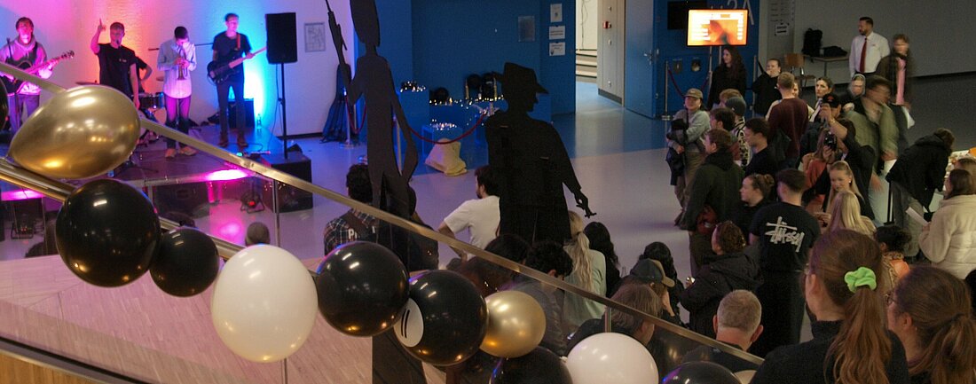 Auf dem Bild sieht man viele Leute vor dem Hörsaal 3A. Es gibt eine Band, viele Leute auf der Treppe und einen Stand mit Merch und Aktionen.