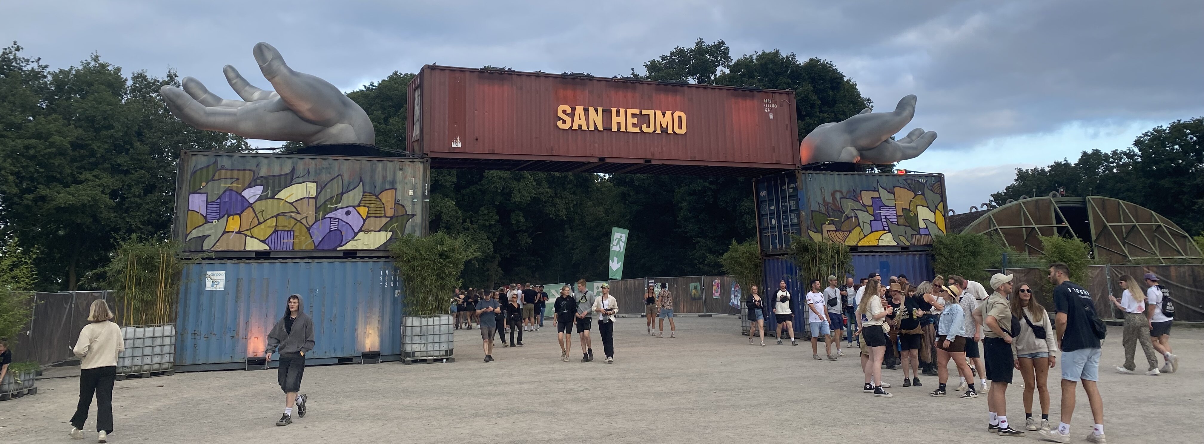 Auf dem Foto ist der Eingang von San Hejmo zu sehen. Auf einem roten Container steht groß San Hejmo. Rechts und links von dem Container sind zwei silberne Hände. Vor diesem Eingang laufen Besucher:innen umher. 