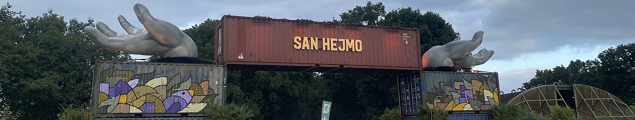 Auf dem Foto ist der Eingang von San Hejmo zu sehen. Auf einem roten Container steht groß San Hejmo. Rechts und links von dem Container sind zwei silberne Hände. Vor diesem Eingang laufen Besucher:innen umher.