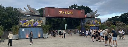 Auf dem Foto ist der Eingang von San Hejmo zu sehen. Auf einem roten Container steht groß San Hejmo. Rechts und links von dem Container sind zwei silberne Hände. Vor diesem Eingang laufen Besucher:innen umher. 