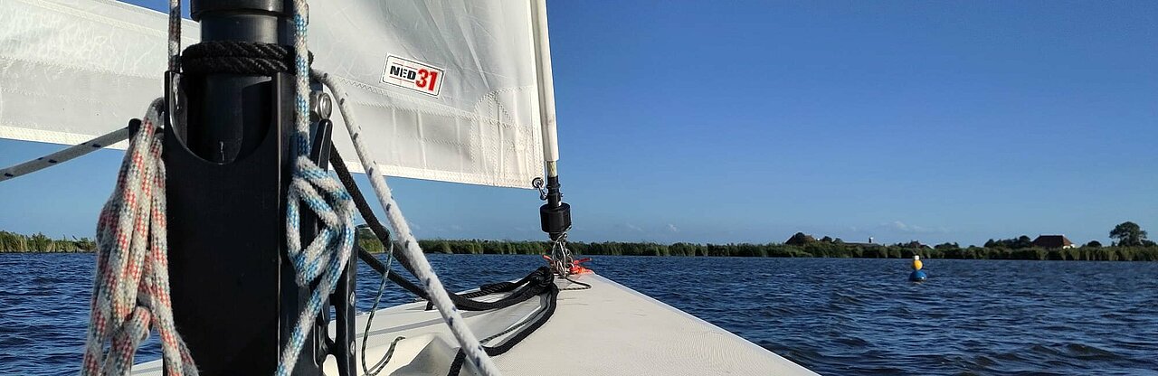 Auf dem Bild zu sehen ist der Bug eines kleinen Segelboots und ein Teil des Masts mit Segel und Leinen