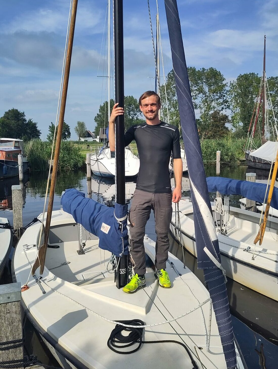 Auf dem Bild ist Max Oehler stehend auf dem Deck eines Segelboots zu sehen, das in einem Hafen liegt.