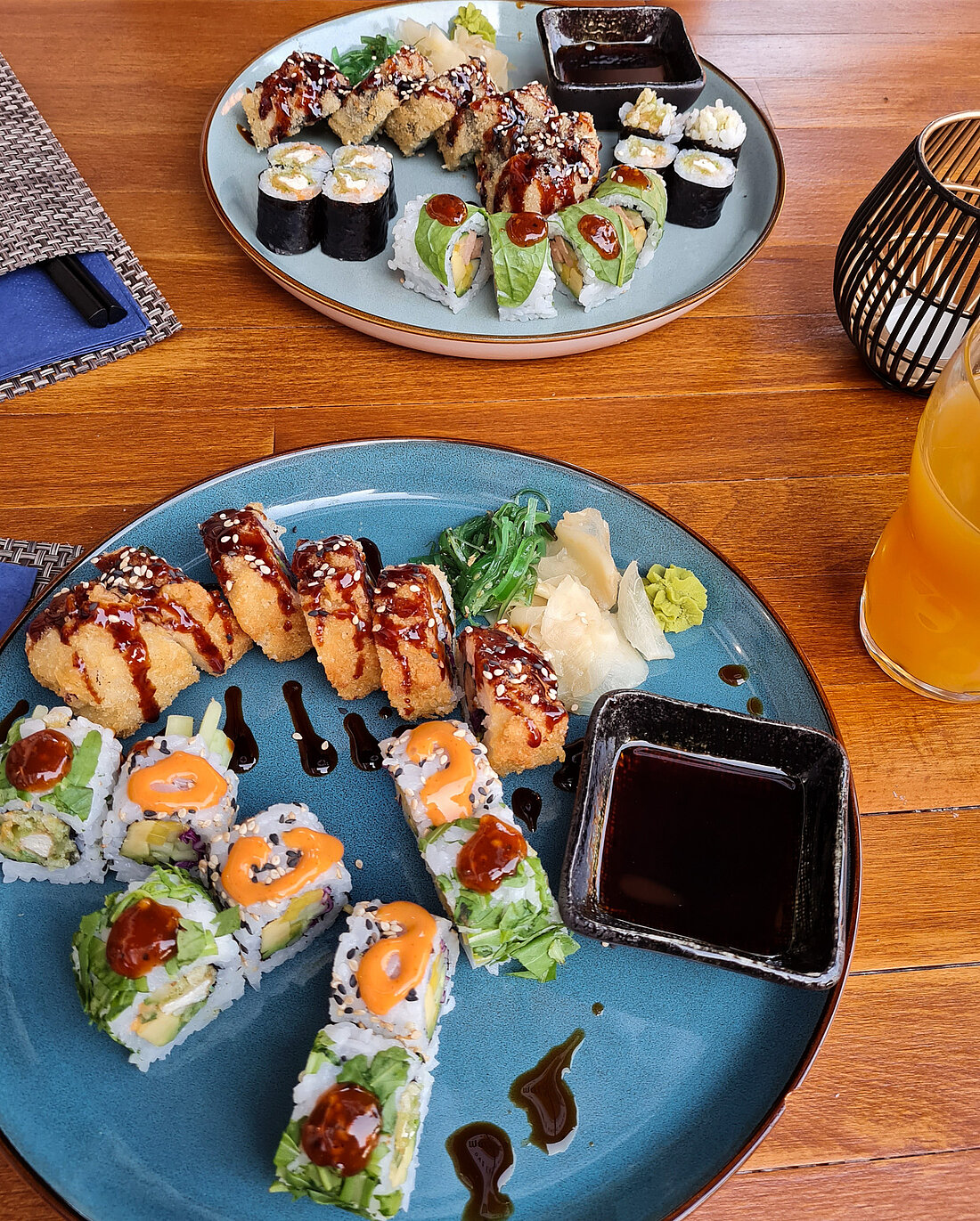 Auf dem Foto sind zwei Teller mit unterschiedlich zubereiteten Sushi Rollen und eine Schale mit Sojasauce. Daneben ein Glas Saft.