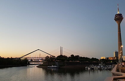 Auf dem Bild ist der Hafen zu sehen. Rechts am Bildrand ist der Rheinturm. Im Hintergrund geht gerade die Sonne unter.