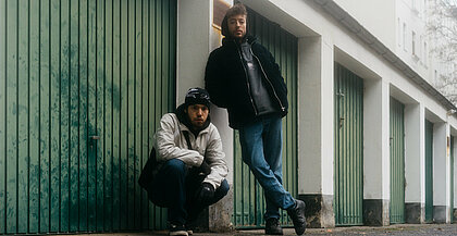 Zwei Männer posieren vor einem weißen Garagengebäude mit grünen Toren. Der linke Mann hockt, der rechte Mann steht aufrecht daneben. Beide schauen in die Kamera.