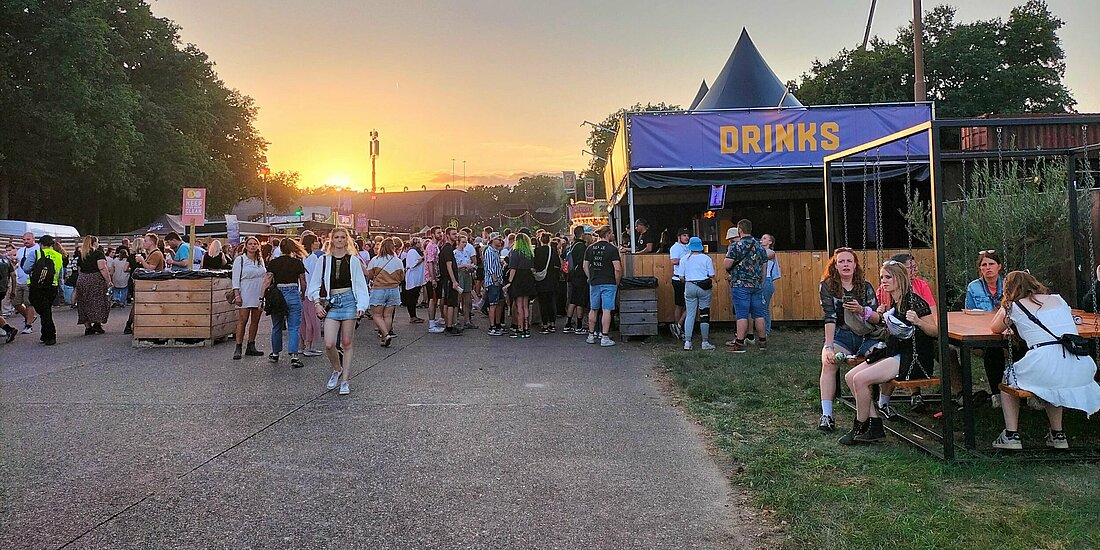 Auf dem Foto ist eine Fußgängerzone zu sehen auf der viele Leute entlang laufen. Rechts ist ein blaues Zelt mit der Aufschrift Drinks. Die Sonne geht gerade unter.
