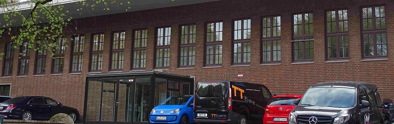 Auf dem Foto ist das ZDM zu sehen. Es ist ein längliches Backsteingebäude vor dem Autos stehen. 