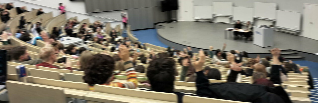 Auf dem Bild sind Studierenden in einem Hörsaal zu sehen. Sie stimmen ab und heben dabei die Hände.