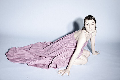 Zu sehen ist die Sängerin Novaa in einem rosanen Kleid, wie sie auf dem Boden kniet und in die Kamera blickt