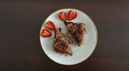 Auf dem Bild sind zwei Nussecken auf einem weißen Teller mit goldenem Rand zu sehen. Zusätzlich liegen dort zwei aufgeschnittene Erdbeeren in Herzform.