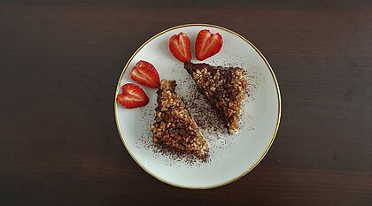 Durch zwei Schnitte lassen sich Erdbeeren ganz leicht zu Herzen formen Auf dem Bild sind zwei Nussecken auf einem weißen Teller mit goldenem Rand zu sehen. Zusätzlich liegen dort zwei aufgeschnittene Erdbeeren in Herzform.