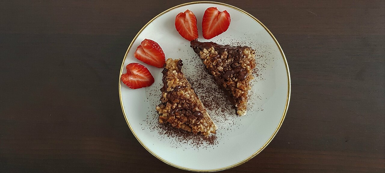 Durch zwei Schnitte lassen sich Erdbeeren ganz leicht zu Herzen formen Auf dem Bild sind zwei Nussecken auf einem weißen Teller mit goldenem Rand zu sehen. Zusätzlich liegen dort zwei aufgeschnittene Erdbeeren in Herzform.