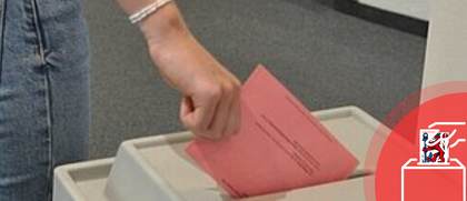 Auf dem Foto ist eine Wahlurne zu sehen. Jemand wirft gerade einen roten Wahlzettel ein.