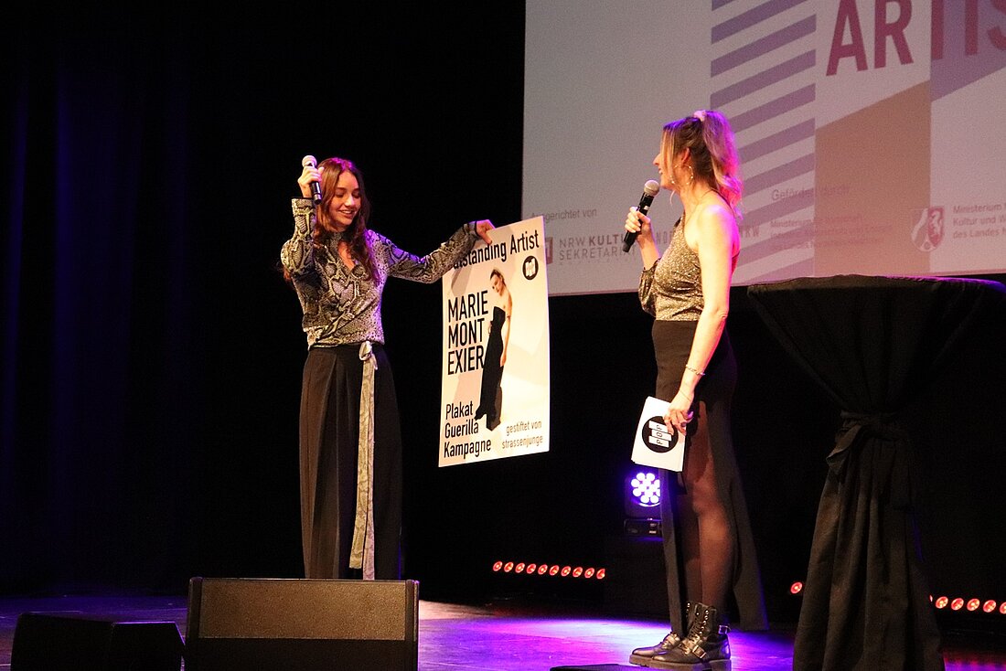 Zu sehen ist die DJ Marie Montexier mit der Moderatorin Linda Reitinger auf der Bühne. Montexier erhielt soeben ihren Preis und hält ein weißes Pappschild in den Händen, auf dem ihr Name steht.