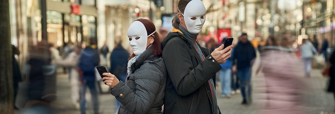 Zwei Menschen stehen in der Fußgängerzone und sind in ihr Handy vertieft. Weiße Masken symbolisiern dass sie sich durch ihre Handys von ihrem Umfeld abschotten.