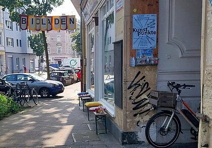 Auf den Bild ist ein Bürgersteig zu sehen, links parkenden Autos, rechts stehen vor einen Gründerzeitgebäude zwei Bierzeltbänke, auf denen bunte Kissen liegen. Zwischen den Gebäude und einen Baum ist eine Girlande gespannt, auf der „Bioladen“ steht. 