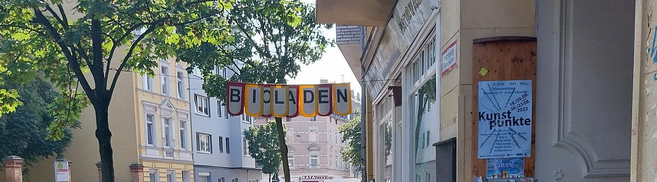 Auf den Bild ist ein Bürgersteig zu sehen, links parkenden Autos, rechts stehen vor einen Gründerzeitgebäude zwei Bierzeltbänke, auf denen bunte Kissen liegen. Zwischen den Gebäude und einen Baum ist eine Girlande gespannt, auf der „Bioladen“ steht. 