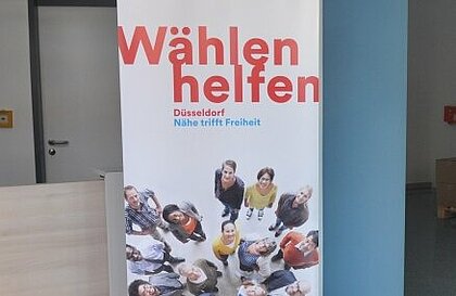 Es ist ein Foto vorhanden. Zu sehen ist ein Roll-up auf dem in roter Schrift “Wählen helfen” steht.