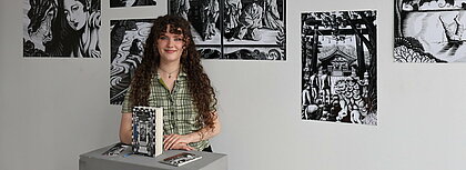 Auf dem Foto ist Ann-Kathrin Peuthen vor ihren Illustrationen zu sehen. Sie lächelt in die Kamera.
