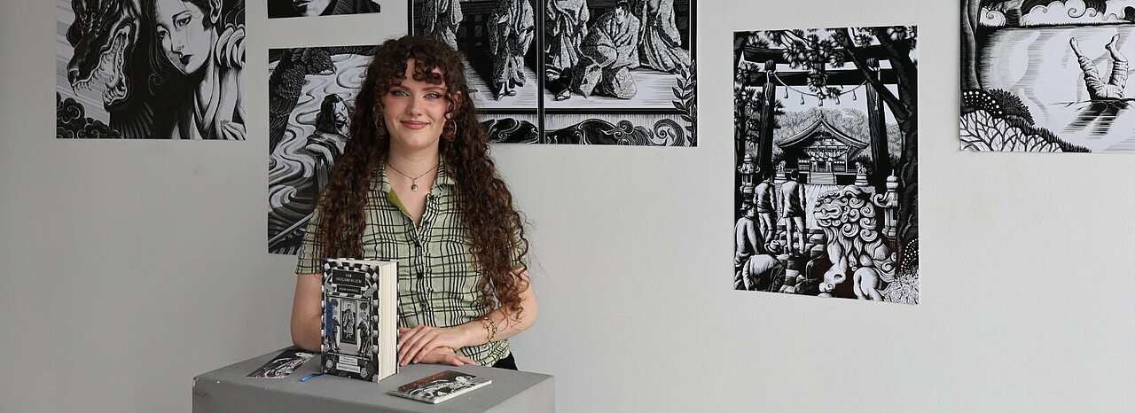 Auf dem Foto ist Ann-Kathrin Peuthen vor ihren Illustrationen zu sehen. Sie lächelt in die Kamera.