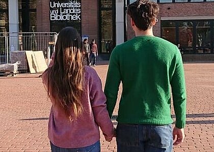 Auf dem Bild sieht man ein junge Frau und einen jungen Mann von hinten. Sie gehen händchenhaltend in der Abendsonne über den Campus der Universität.