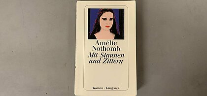Das Cover der deutschen Version von "Mit Staunen und Zittern" Darauf, der Titel und der Name der Autorin, Amelie Nothomb und ein Bild ein Frau vor einem dunkelblauen Hintergrund.