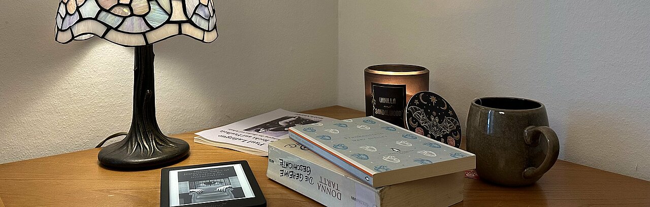 Ein Stapel Bücher liegt unter einer Lampe, daneben stehen eine Duftkerze und eine Tasse Tee.