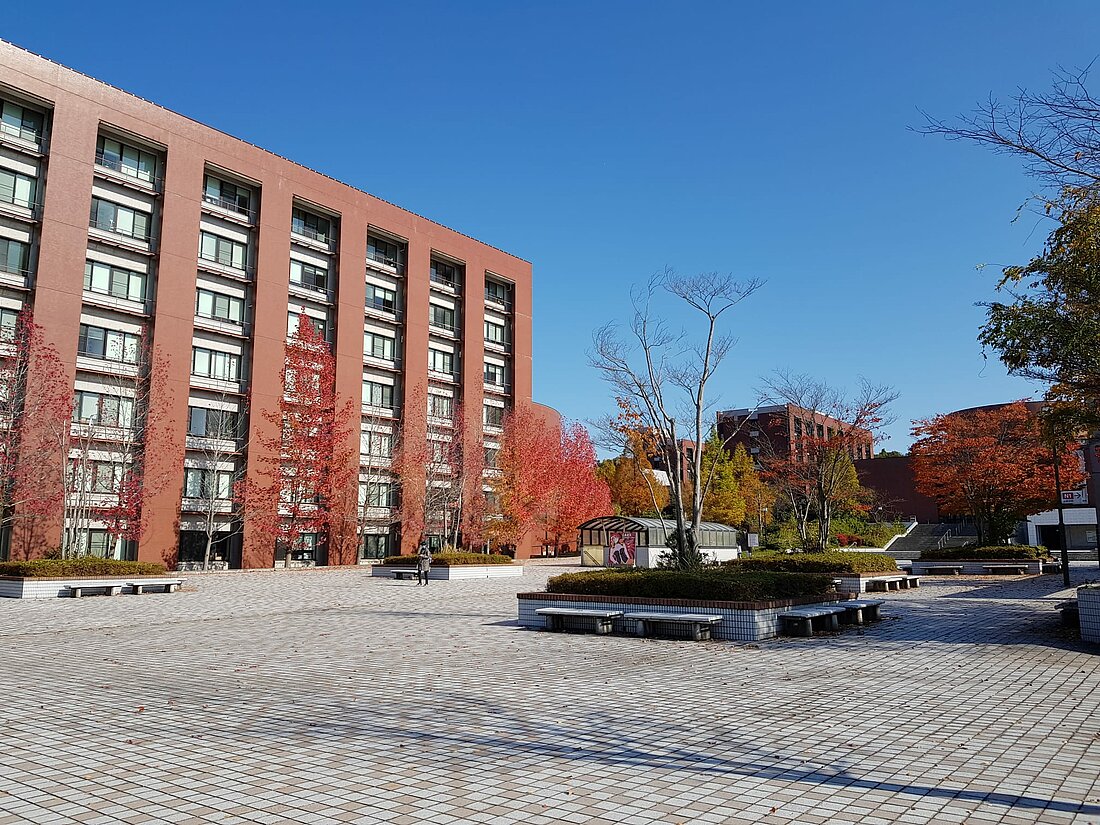 Man sieht einen Ausschnitt des Campus der Kanazawa-Universität. Links ist ein rotes Universitäts-Gebäude unter klarem blauen Himmel, rechts sieht man Pflastersteine und vereinzelnd Bäume und Büsche.