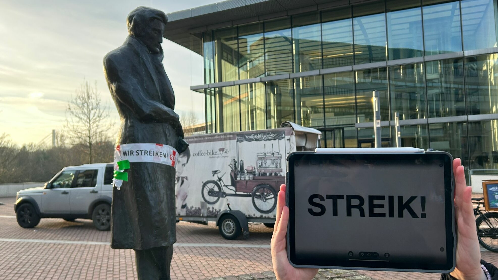 Foto: Lena Sorgalla Zu sehen ist die Heinrich Heine Statue auf dem Campus, welcher ein rotes Band umgebunden wurde. Auf dem Band steht ein Streikaufruf und neben der Statue wird ein IPad mit der Aufschrift „Streik!“ hochgehalten