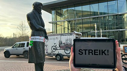 Zu sehen ist die Heinrich Heine Statue auf dem Campus, welcher ein rotes Band umgebunden wurde. Auf dem Band steht ein Streikaufruf und neben der Statue wird ein IPad mit der Aufschrift „Streik!“ hochgehalten