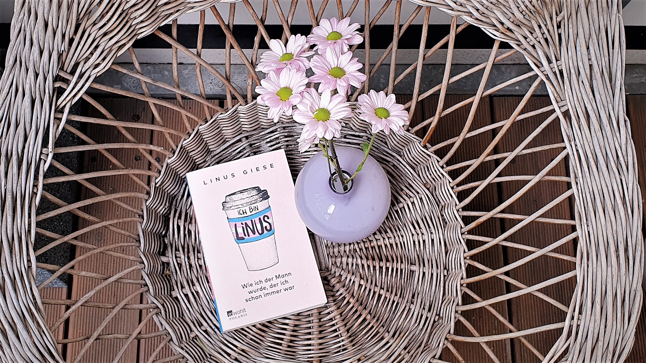 Zu sehen ist das Buch "Ich bin Linus" auf einem Korbstuhl. Rechts vom Buch befindet sich eine lila Blumenvase mit rosa Blumen.
