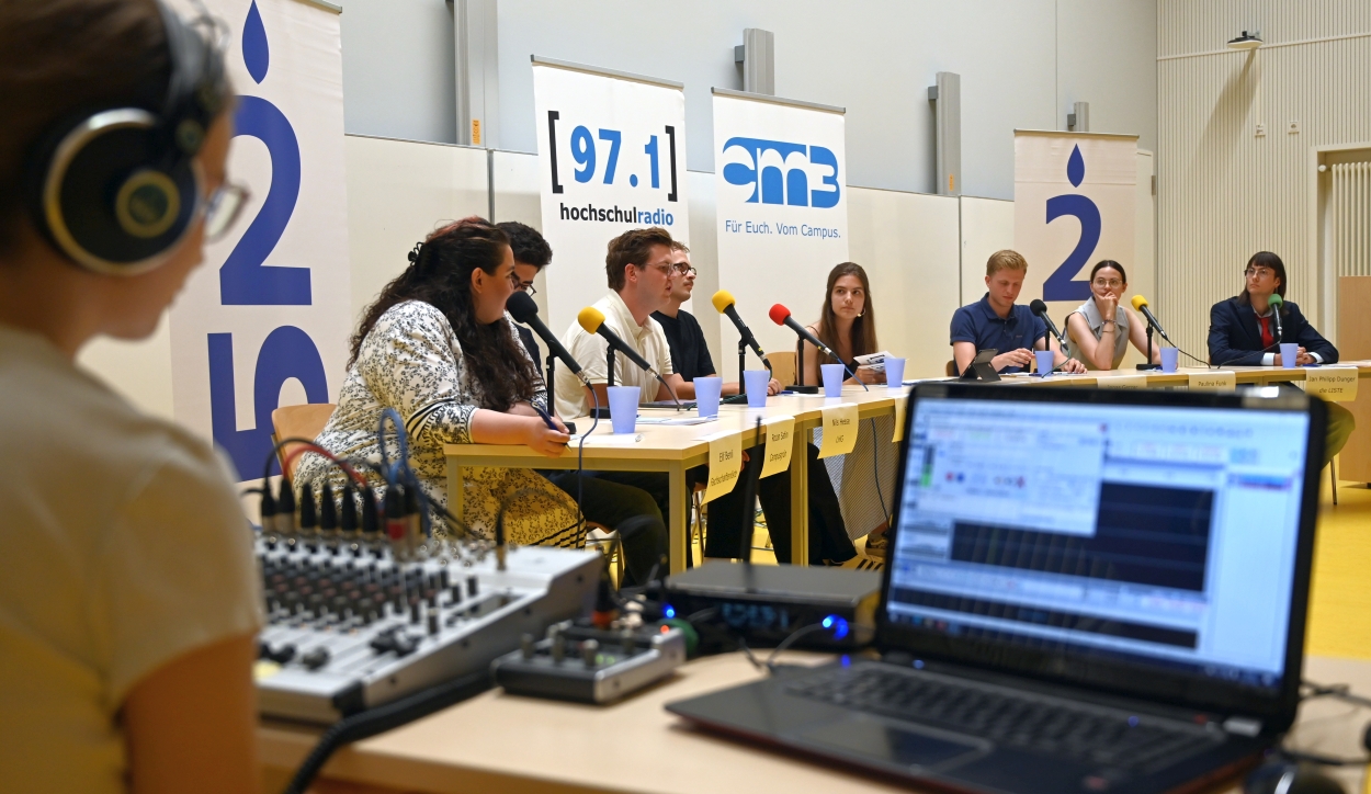 Auf dem Fotos sind die Kandidierenden der Listen und die Moderatorin zu sehen. Sie sitzen im Halbkreis an Tischen. Alle Kandidierenden haben ein Mirofon vor sich stehen. Im Vordergrund sieht man das mobile Aufnahmestudio von hochschulradio düsseldorf, bestehend aus einem Laptop und einem Mischpult.