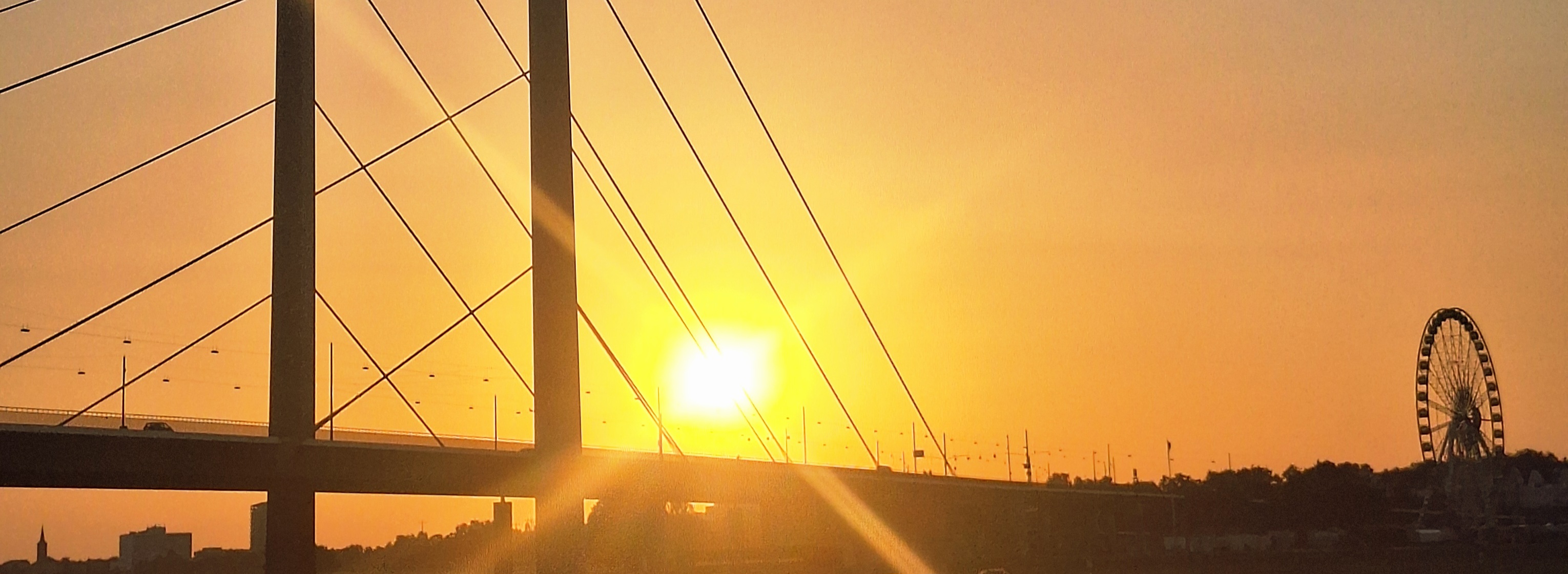 Ein oranger Sonnenuntergang hinter der Rheinkniebrüke mit dem Riesenrad für die Rheinkirmes 