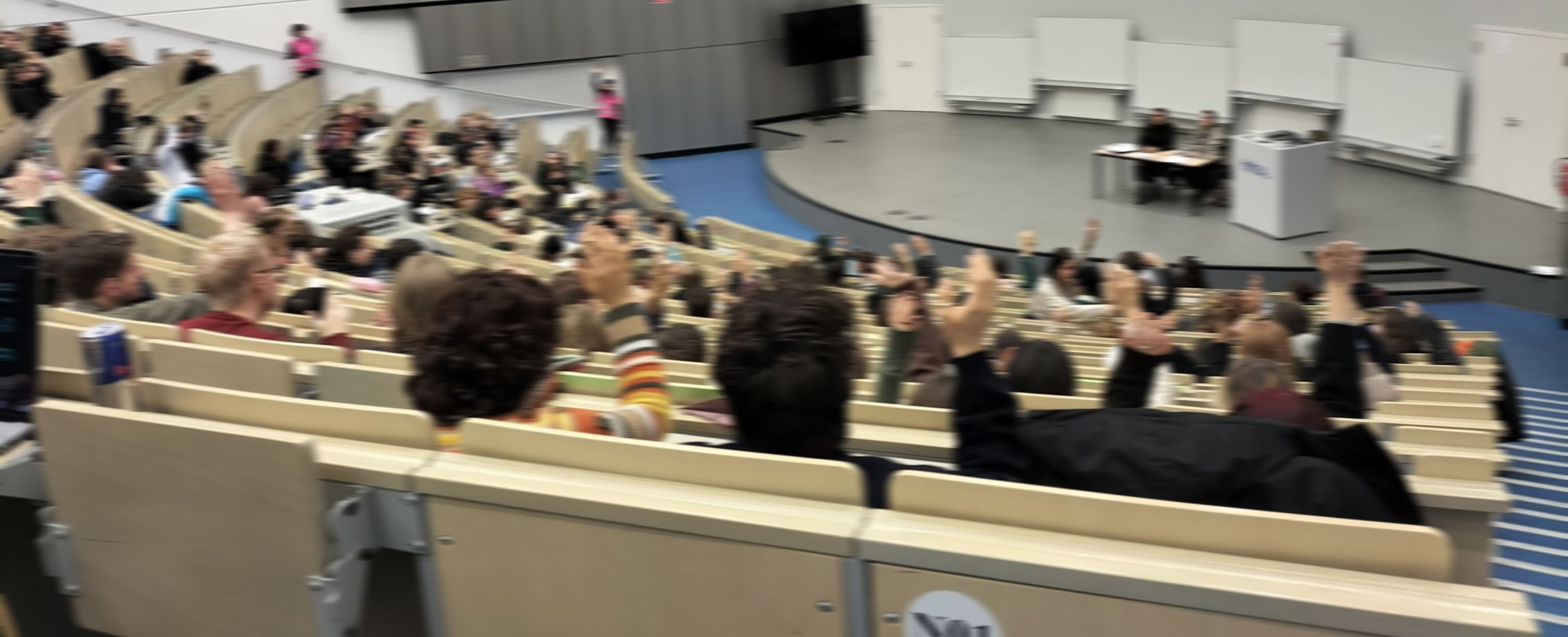 Auf dem Bild sind Studierenden in einem Hörsaal zu sehen. Sie stimmen ab und heben dabei die Hände.